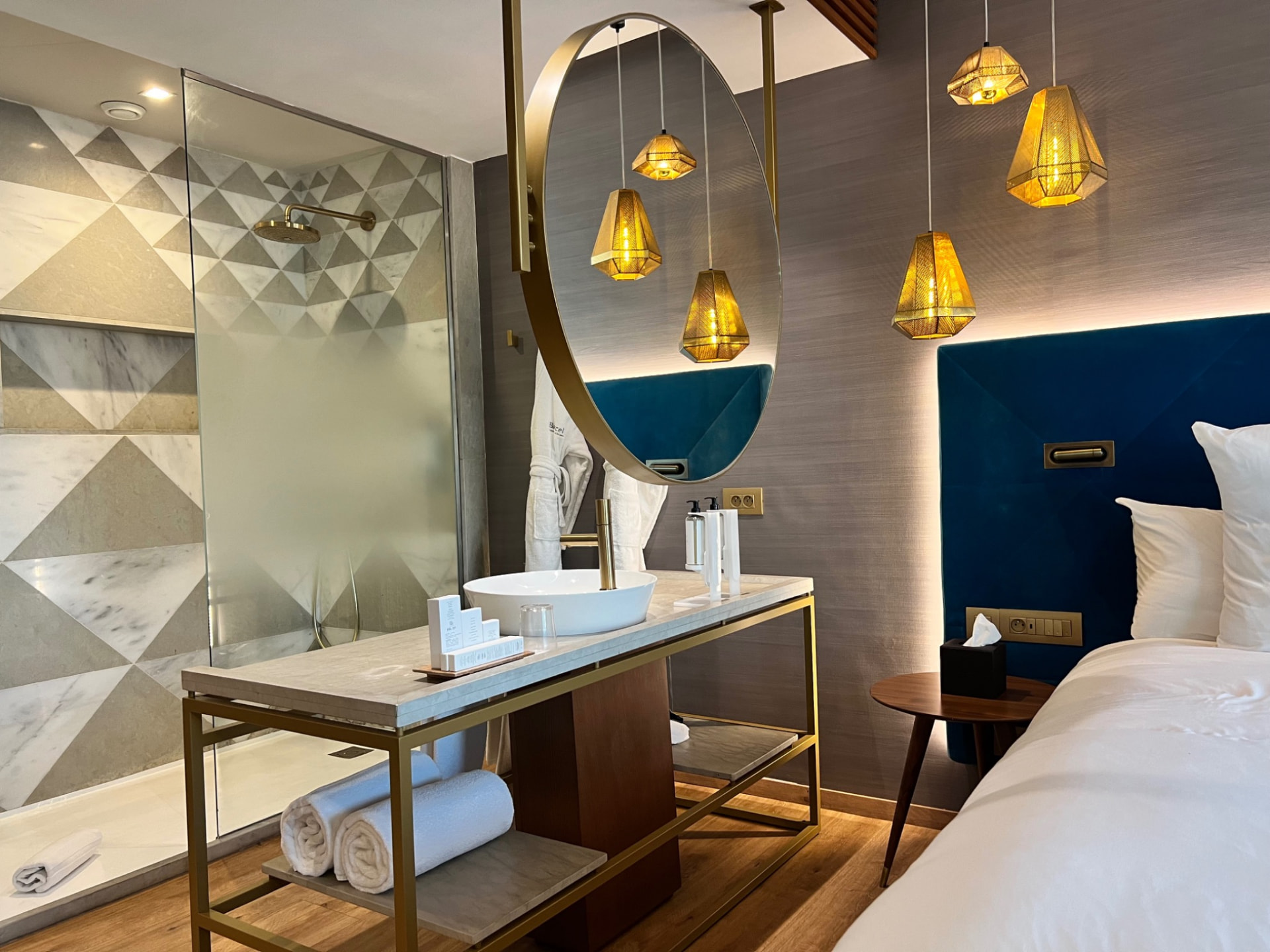 A contemporary Moroccan hotel bedroom and open bathroom with brass fittings and a deep blue velvet headboard