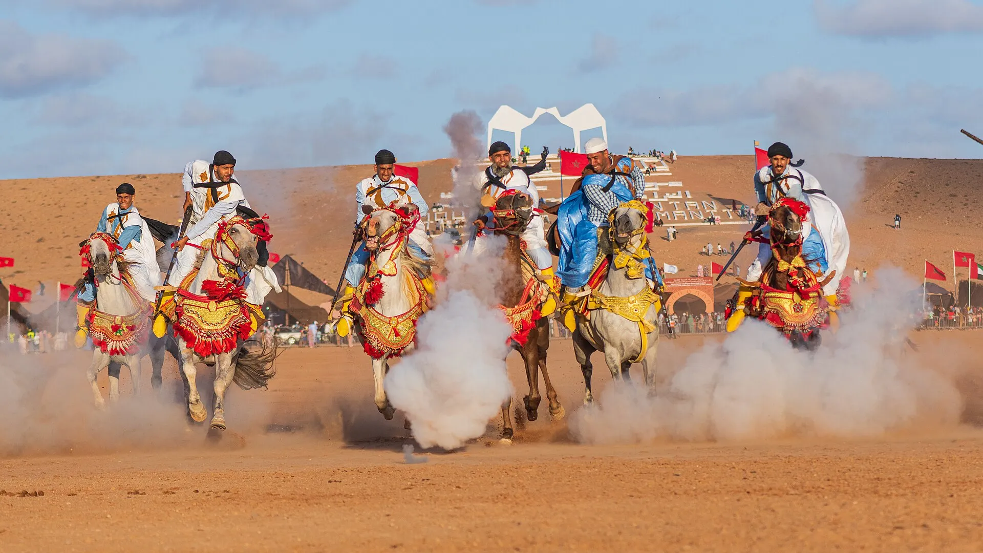Tbourida horsemen galloping through gunpowder smoke at the Moussem of Tan-Tan, with flags and the festival arena behind