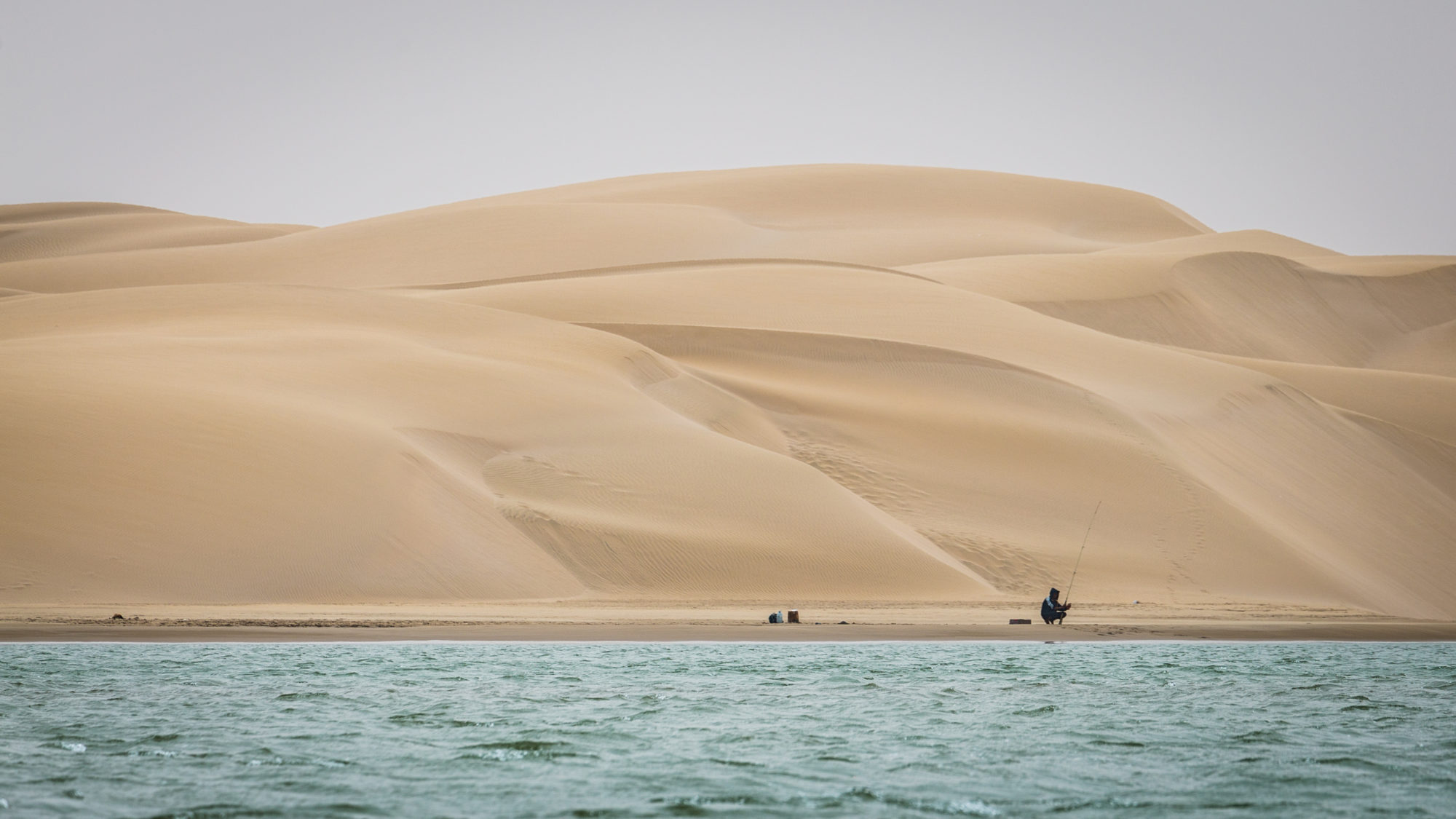 A lone fisherman seated on the shoreline below huge dunes at Khnifiss lagoon