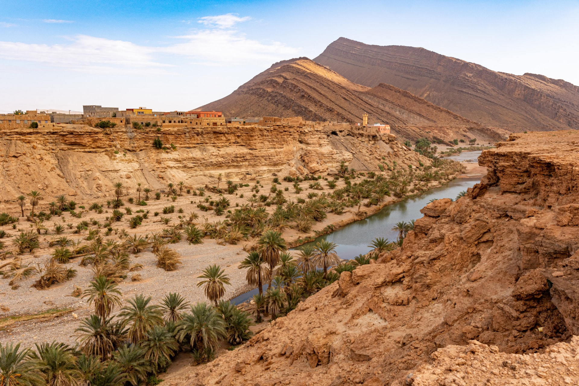 A palm-lined river valley and village below a steep slanted mountain ridge in Morocco's Anti-Atlas