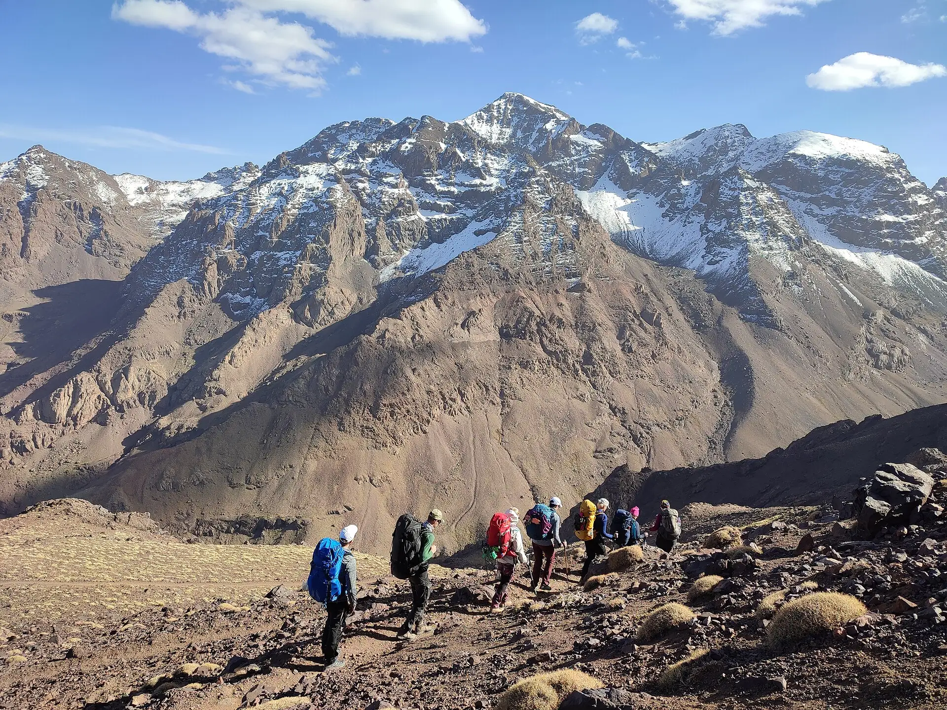 Trekkers walking a stony trail below the snow-streaked peaks of the Toubkal massif in the Moroccan High Atlas