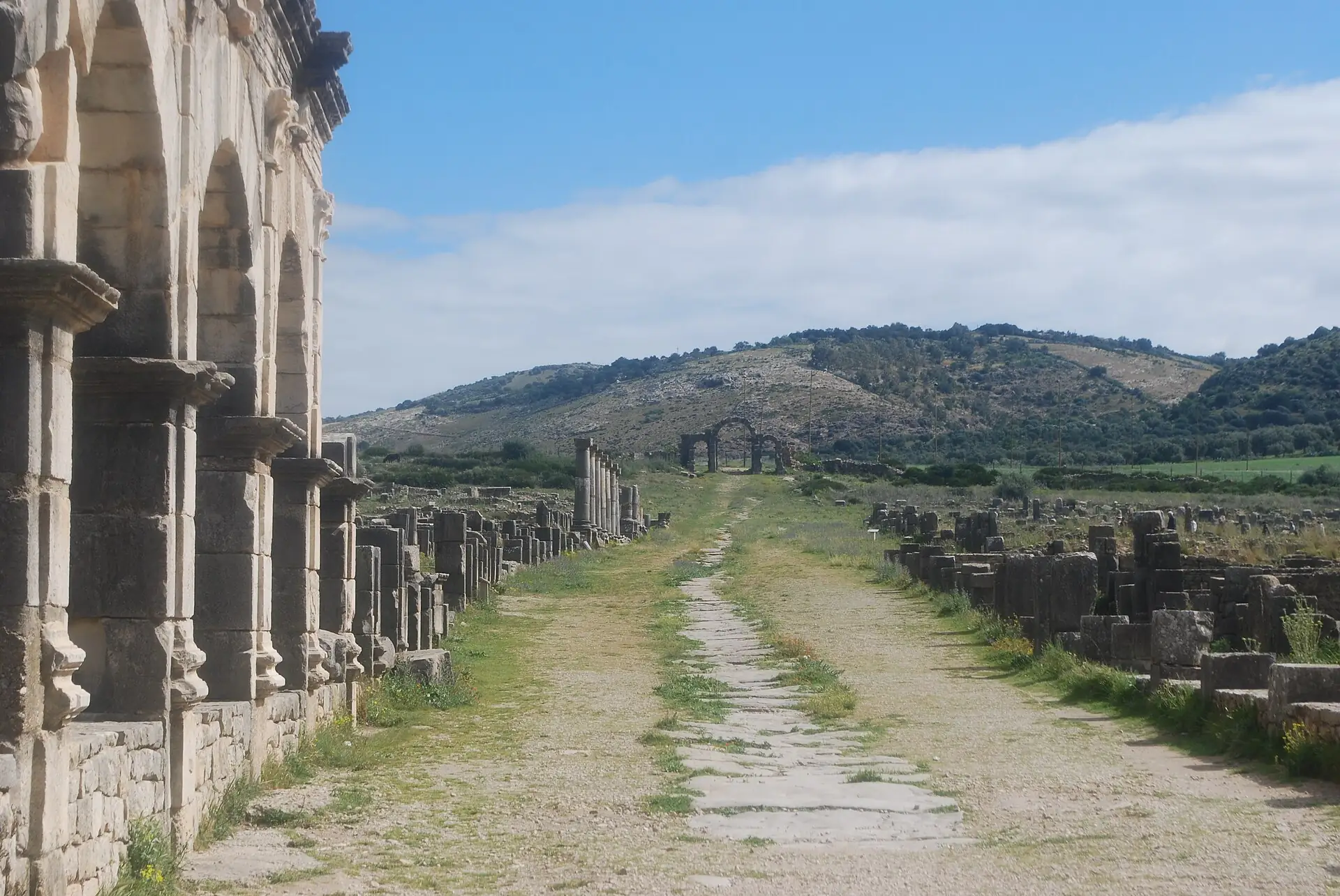 The colonnaded main street of Volubilis leading past the basilica towards the triumphal arch under a bright blue sky